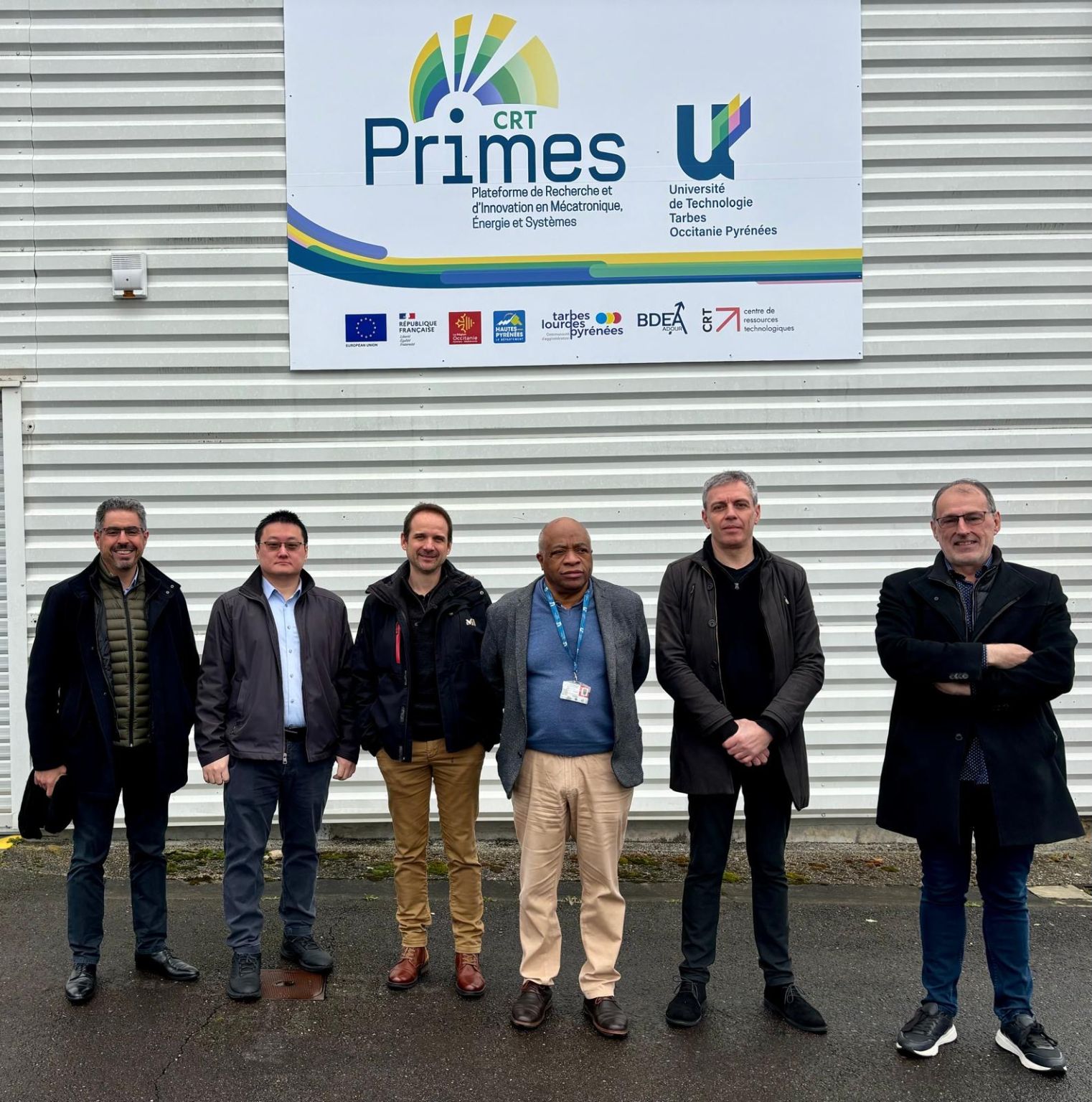 6 hommes posant devant une facade avec un panneau CRT primes