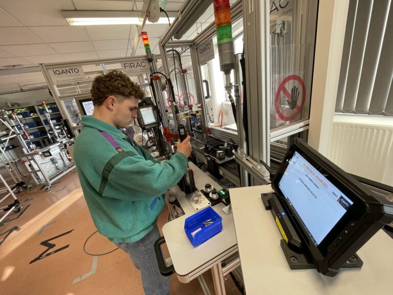 L'image montre un jeune homme travaillant dans un atelier industriel moderne, un laboratoire technologique ou une salle de formation technique. Il est debout devant un poste de travail automatisé et semble manipuler un outil ou une pièce mécanique.