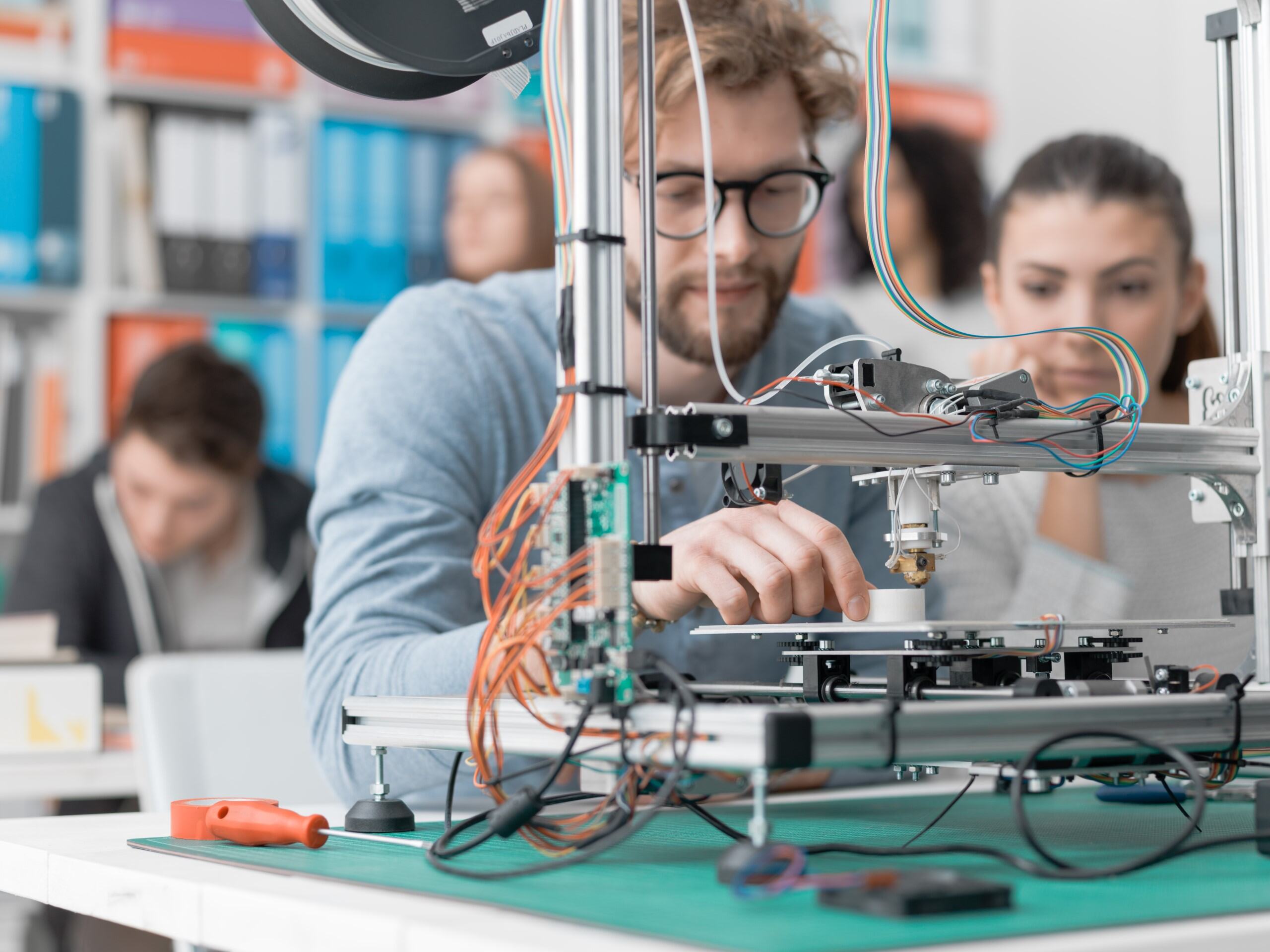 L'image montre deux jeunes adultes concentrés sur le réglage ou l'observation d'une imprimante 3D. La scène se déroule dans un espace de travail collaboratif, un FabLab ou une salle de classe, avec d'autres personnes floues à l'arrière-plan.