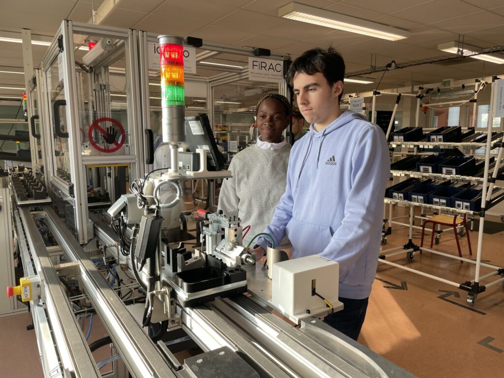 L'image montre deux jeunes étudiants travaillant sur une ligne de convoyage industrielle automatisée. Ils observent le fonctionnement des mécanismes installés sur le rail.