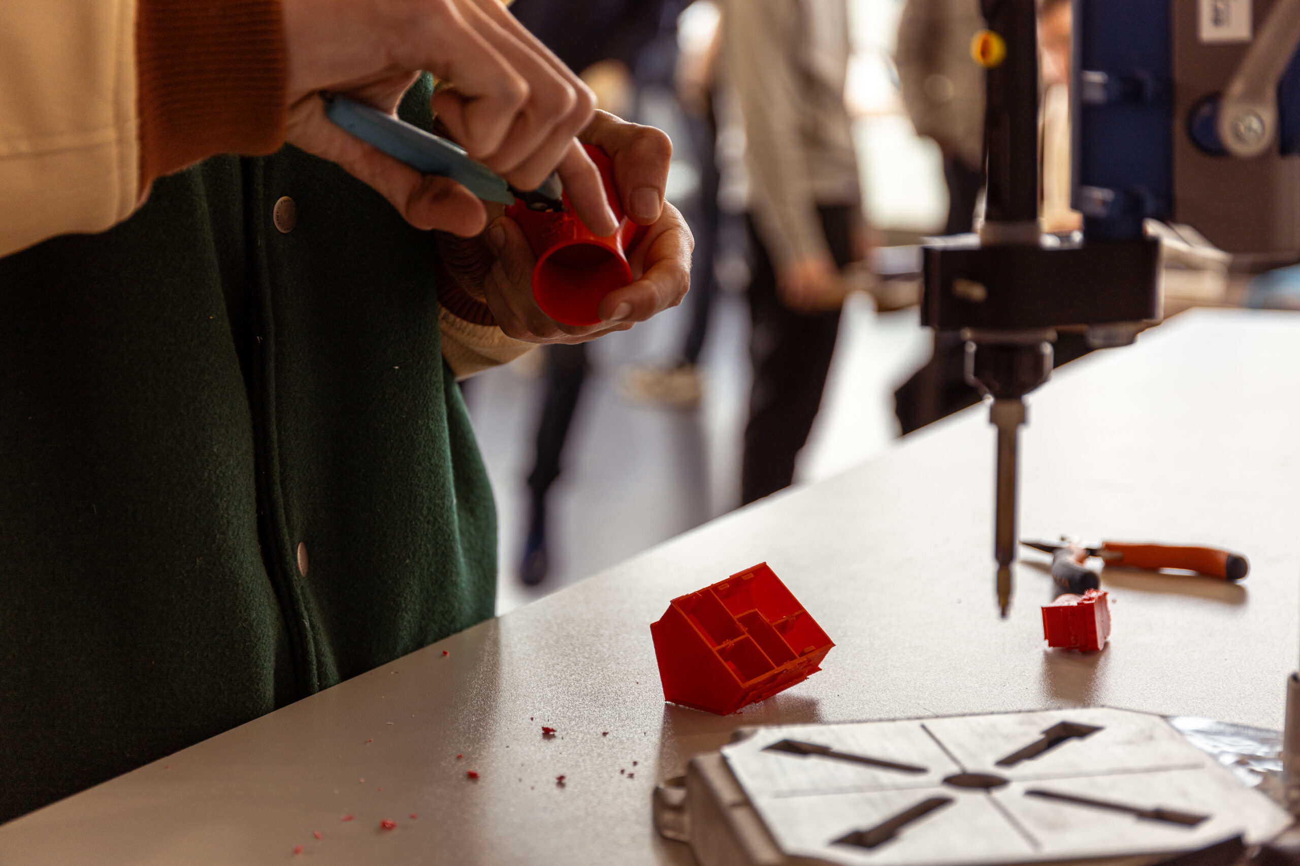 gros plan montre les mains d'une personne en train de travailler minutieusement sur une petite pièce en plastique rouge à l'aide d'une pince, dans un atelier de fabrication ou un FabLab. L'arrière-plan est flou, mettant l'accent sur le travail manuel.