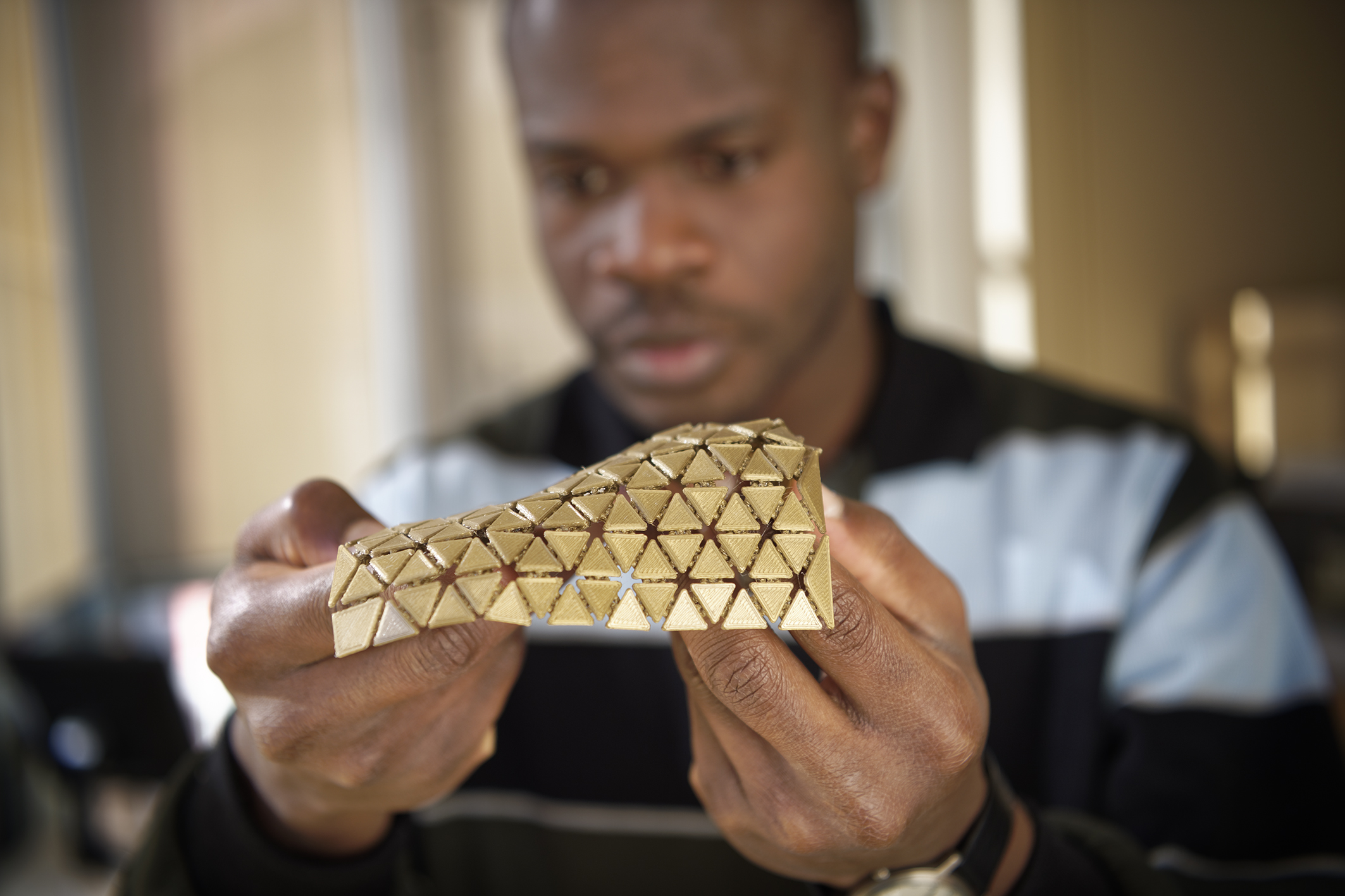 Un homme regarde attentivement et manipule avec ses deux mains un matériau flexible de couleur dorée, composé d'une multitude de petits triangles articulés en relief.