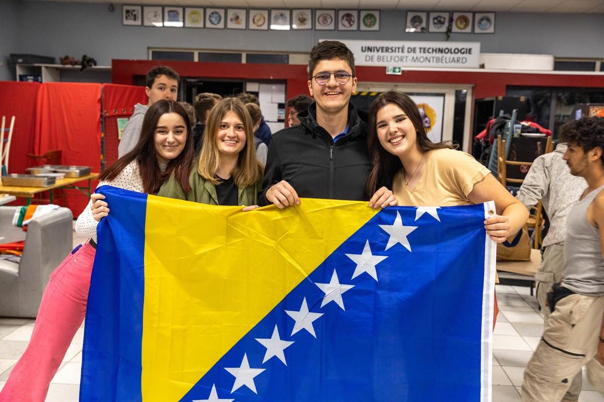 Quatre étudiants souriants posent ensemble en intérieur en tenant le drapeau de la Bosnie-Herzégovine au premier plan. En arrière-plan, on aperçoit d'autres personnes et un panneau indiquant « Université de Technologie de Belfort-Montbéliard ».