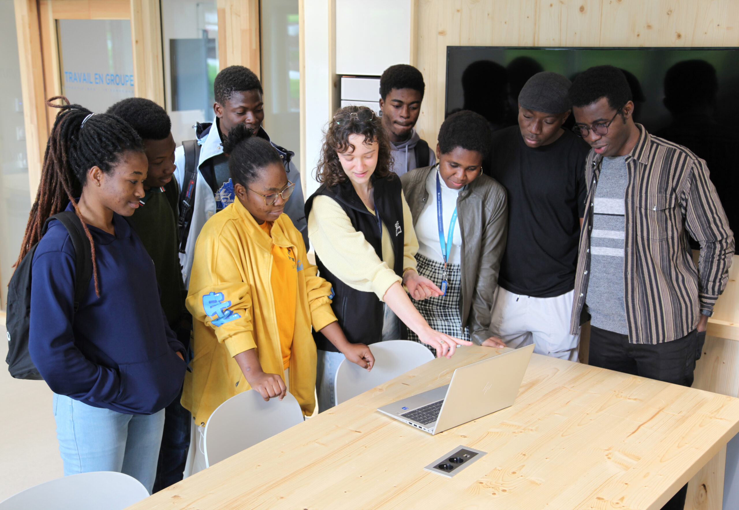 Un groupe d'étudiants debout, derrière un ordinateur portable. Une femme montre l'écran avec son doigt.