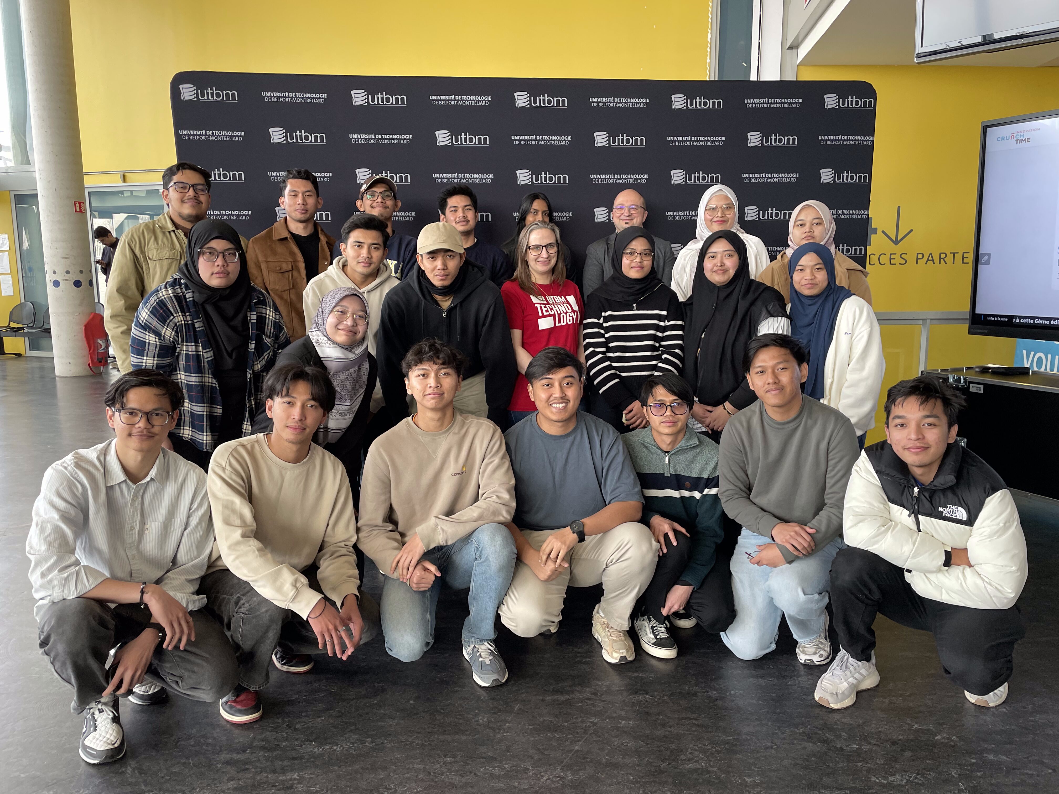 Groupe d'étudiants malaysiens devant un photocall UTBM dans le hall de l'Axone de Montbéliard