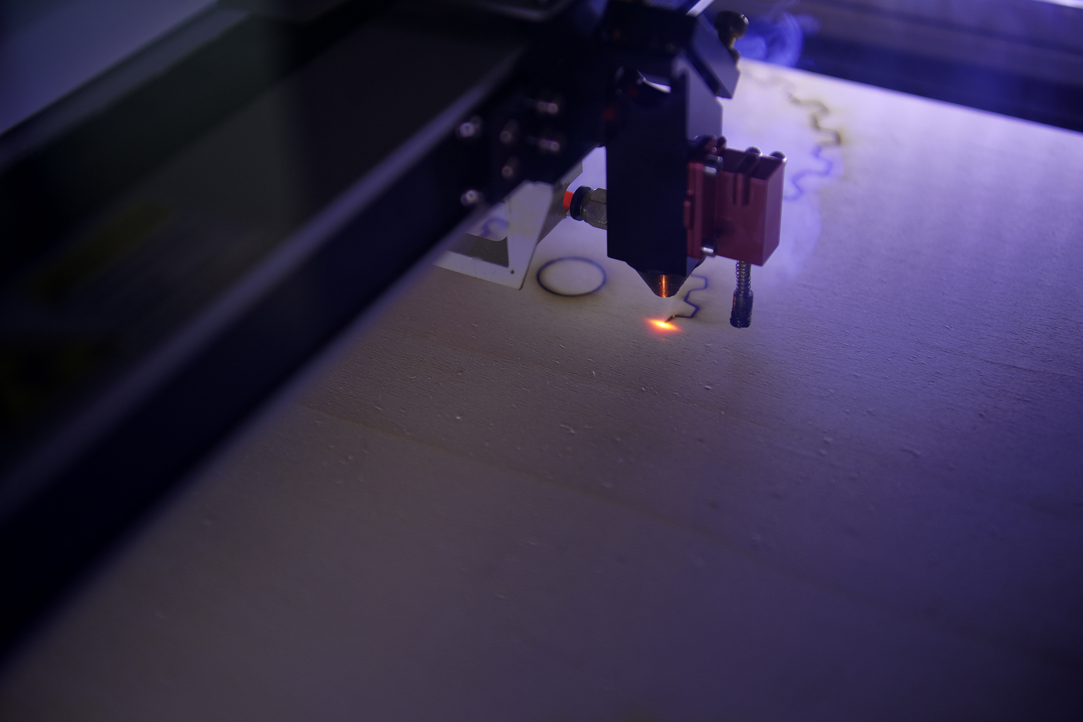 Vue rapprochée d'une tête de découpe laser en action sur une plaque de bois clair sous un éclairage bleuté. Un point lumineux orange vif marque l'endroit exact où le laser découpe des formes géométriques et des lignes sinueuses, dégageant une légère fumée.