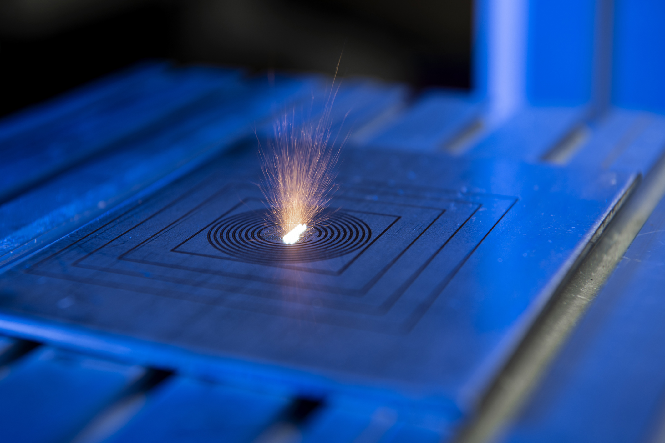 Gros plan sur un processus d'impression 3D métal par fusion laser. Un faisceau lumineux intense frappe une plaque métallique bleutée, générant une gerbe d'étincelles orange au centre de motifs concentriques gravés en forme de spirales et de carrés.