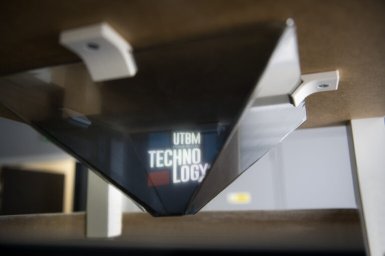 Gros plan en contre-plongée sur un dispositif d'affichage holographique en forme de pyramide inversée, fixé sous un meuble en bois. Au centre du prisme transparent, le logo lumineux « UTBM POWERED BY TECHNOLOGY » apparaît en relief.