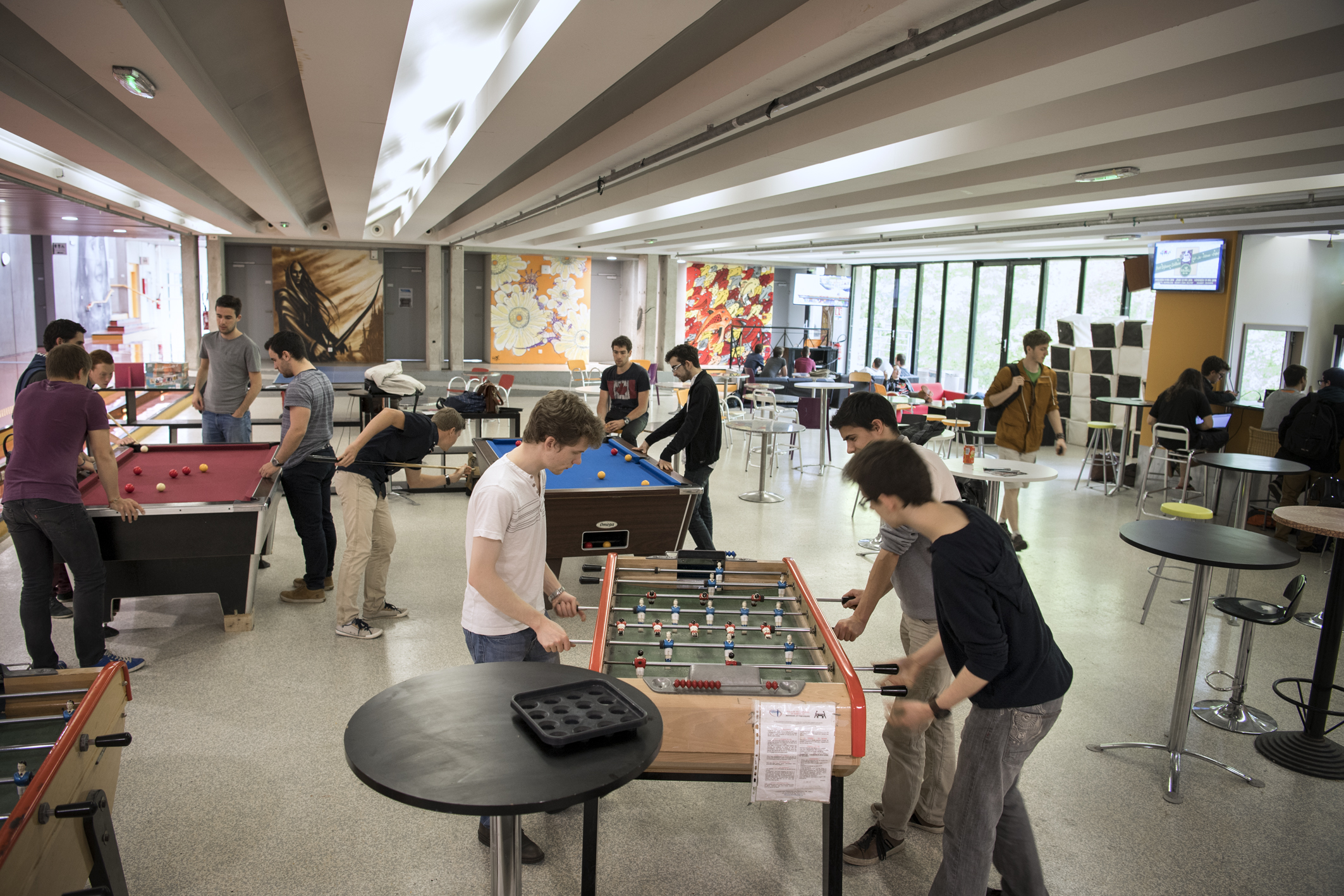 Plusieurs groupes d'étudiants dans une salle de repos. Un groupe joue au baby-foot, un autre groupe joue au billard, d'autres étudiants sont assis
