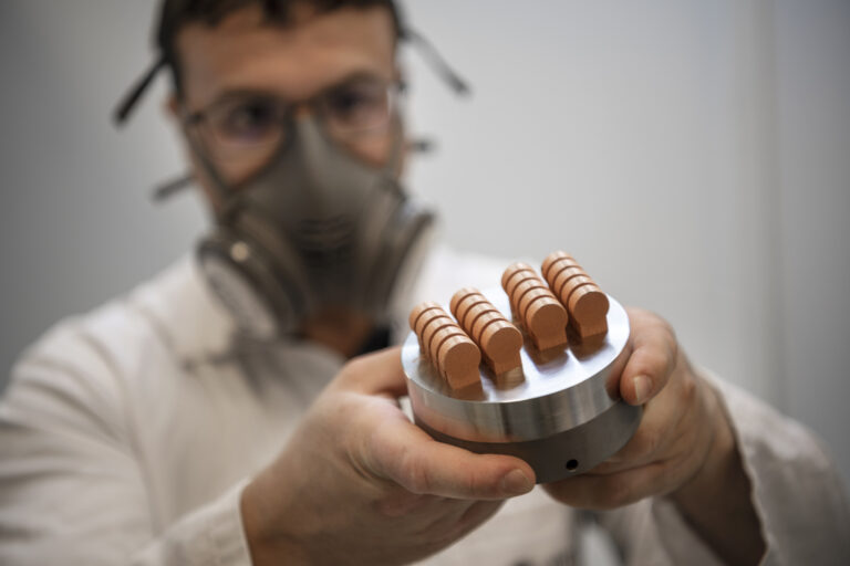 Gros plan sur les mains d'un scientifique en blouse blanche tenant une pièce métallique cylindrique. Sur cette base en acier sont fixées six petites pièces en cuivre identiques, imprimées en 3D et alignées en deux rangées. En arrière-plan flou, le chercheur porte un masque de protection respiratoire.