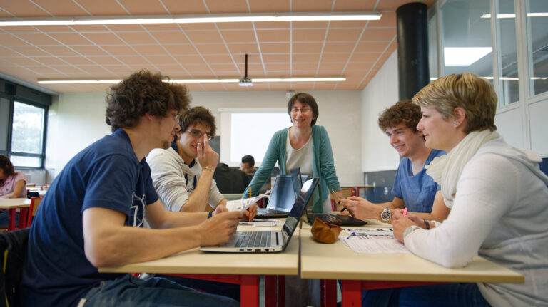 Une enseignante échange avec un groupe de quatre étudiants souriants travaillant ensemble autour d'ordinateurs portables dans une salle de classe.