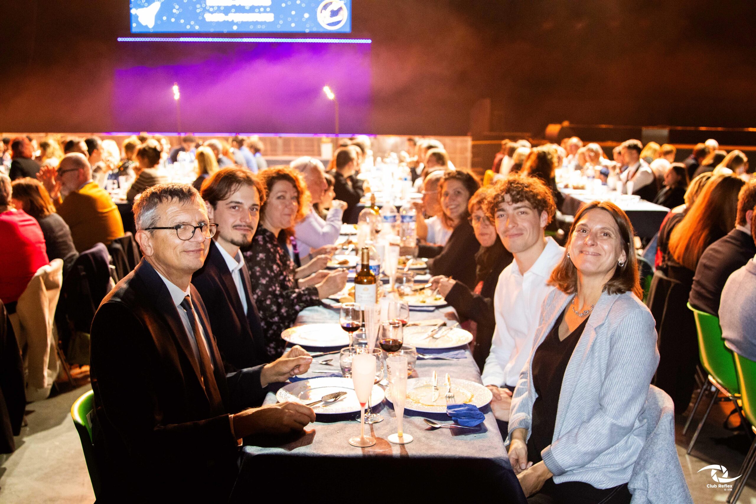 Une grande salle de réception ou de spectacle où de nombreux convives sont attablés pour un dîner de gala officiel. Au premier plan, une tablée d'invités sourit face à l'objectif.