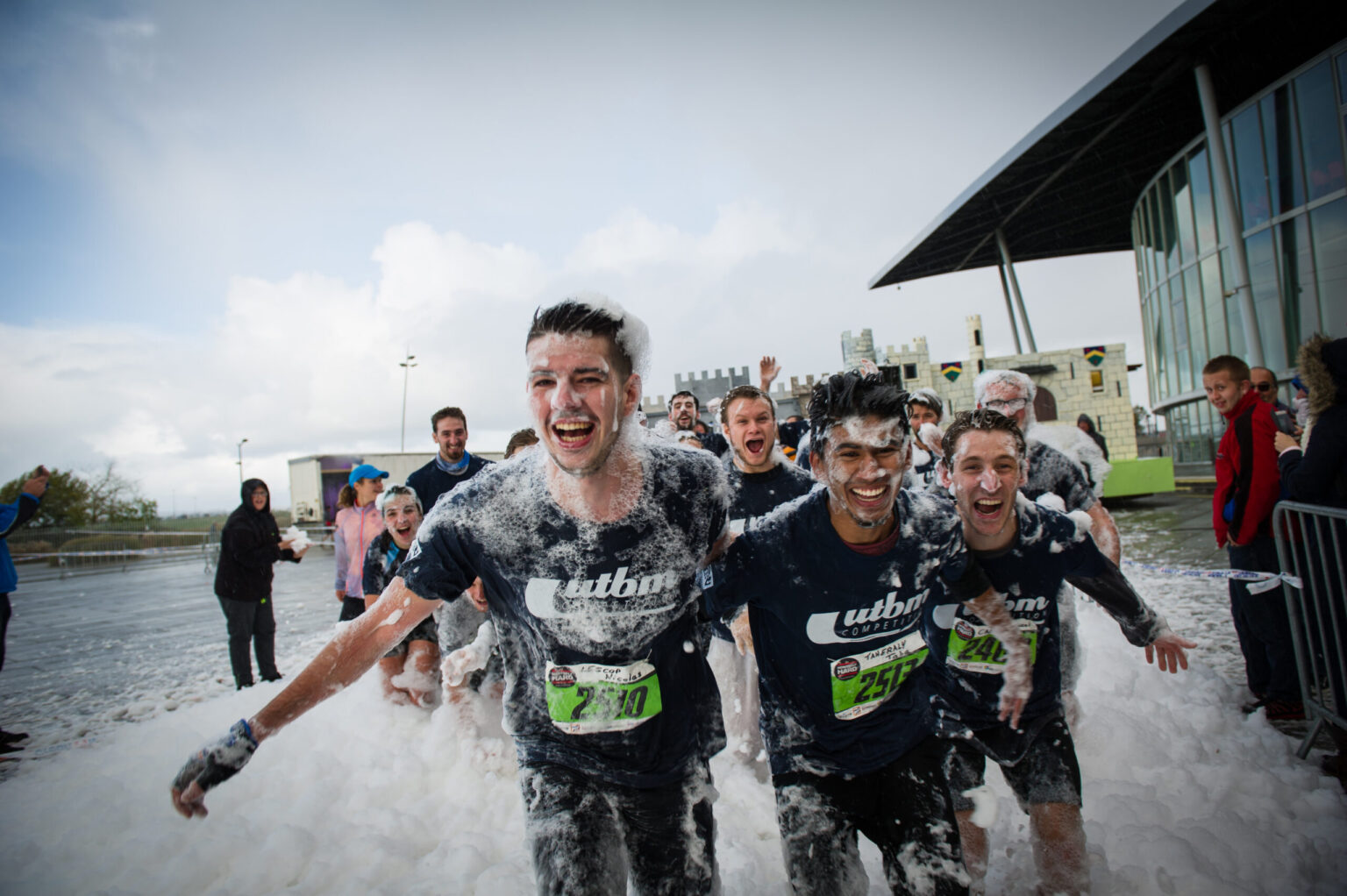 Trois étudiants en t-shirts bleus de l'UTBM courent au milieu d'un épais tapis de mousse blanche lors d'une course festive.