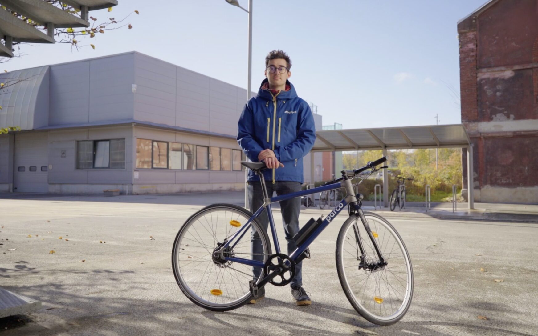 Un jeune homme à lunettes en veste de sport bleue se tient debout en extérieur à côté d'un vélo bleu et gris de la marque Parco.