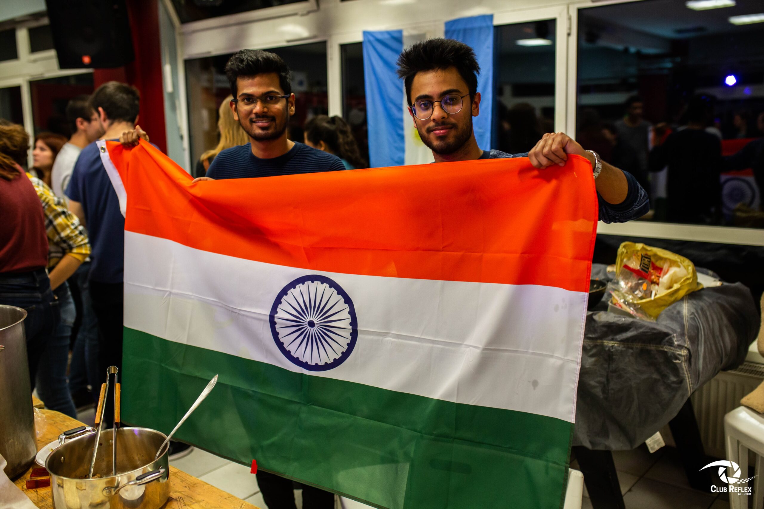 Deux jeunes hommes souriants portent ensemble un grand drapeau de l'Inde lors d'un événement festif ou culturel en intérieur.