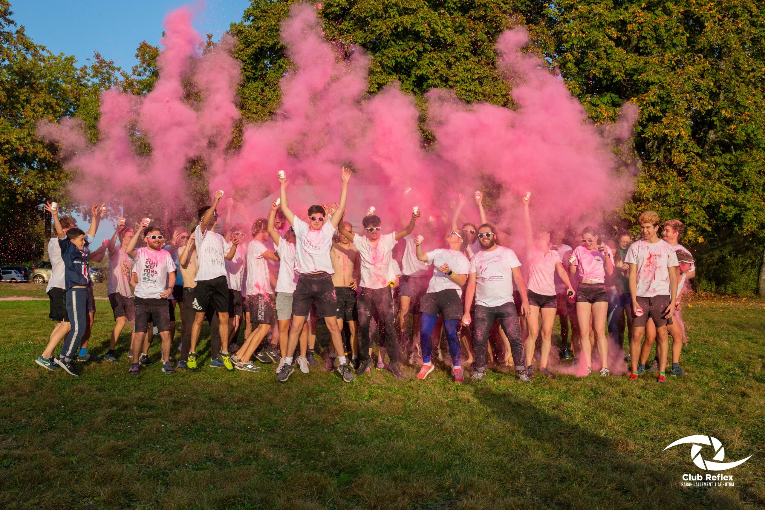Un groupe de jeunes en t shirts blancs célèbrant en extérieur au milieu d'un nuage de poudre rose projeté en l'air, style course Color Run.