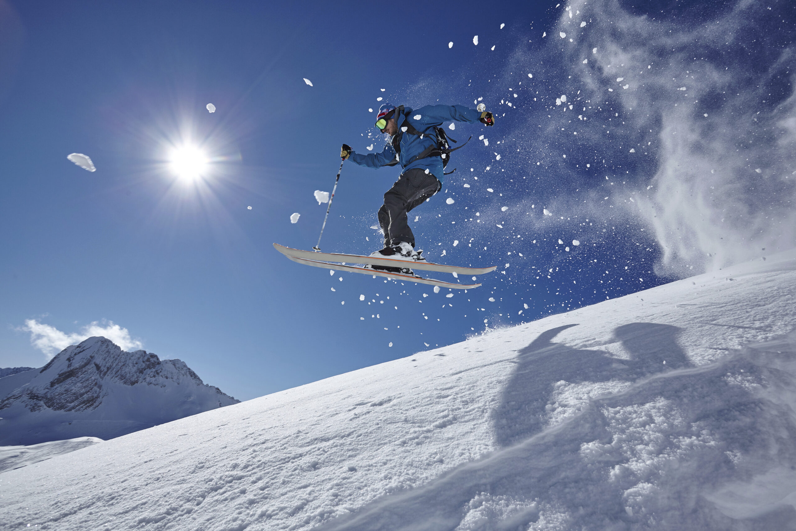 Un skieur en veste bleue réalise un saut dans les airs au-dessus d'une crête enneigée, sous un grand ciel bleu ensoleillé.