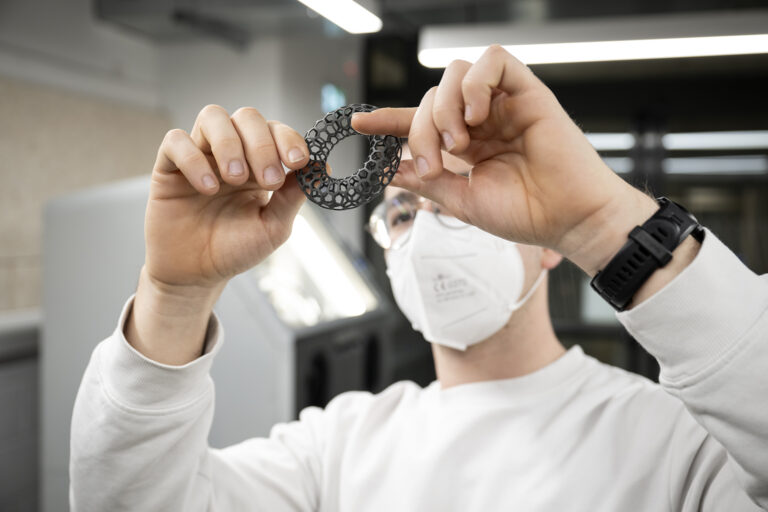 Un ingénieur ou technicien examine de près une pièce circulaire noire, imprimée en 3D avec une structure alvéolaire complexe.