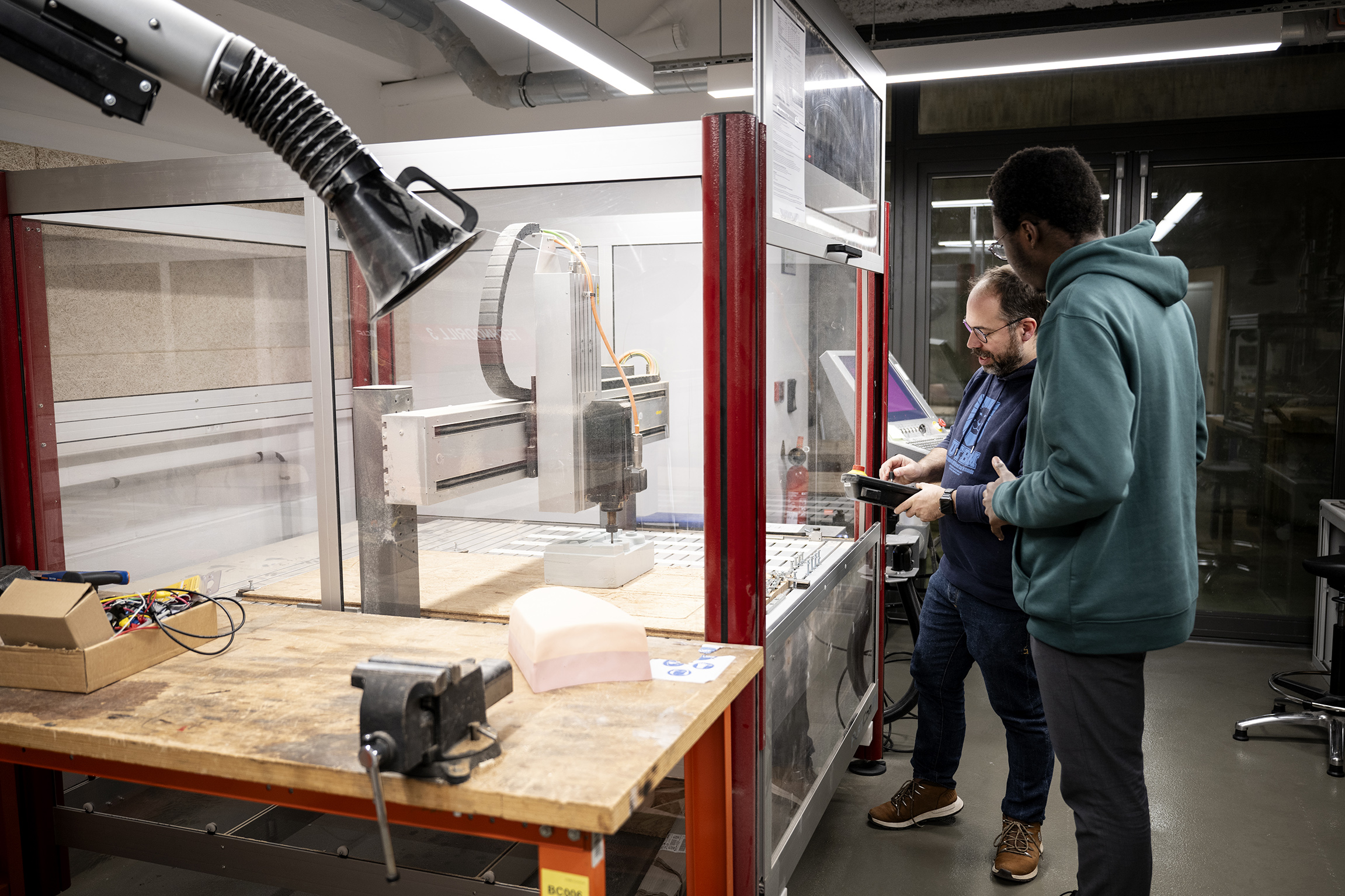 Dans un atelier technologique, un technicien ou enseignant montre le fonctionnement d'une grande fraiseuse numérique protégée par une verrière à un étudiant de dos.
