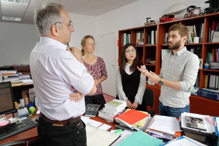 Quatre collègues en discussion autour d’un bureau encombré de documents dans un bureau, lors d’une réunion de travail.