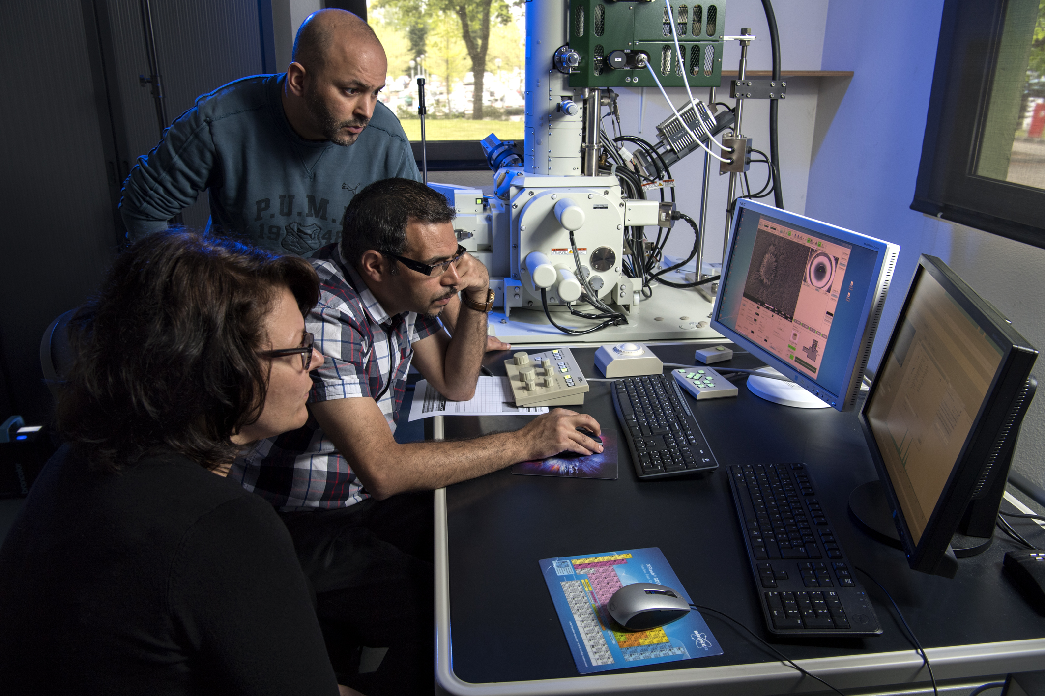 Trois scientifiques, deux hommes et une femme, analysent concentrés des images microscopiques sur des écrans d'ordinateur reliés à un microscope électronique à balayage dans un laboratoire.