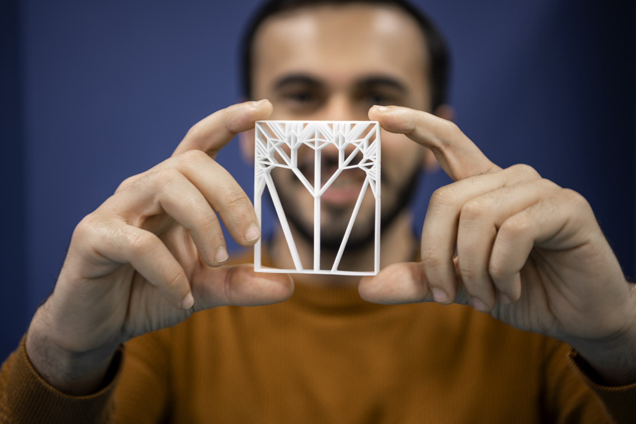 Personne présentant une fine structure blanche imprimée en 3D aux motifs géométriques, tenue entre les doigts devant un fond bleu.